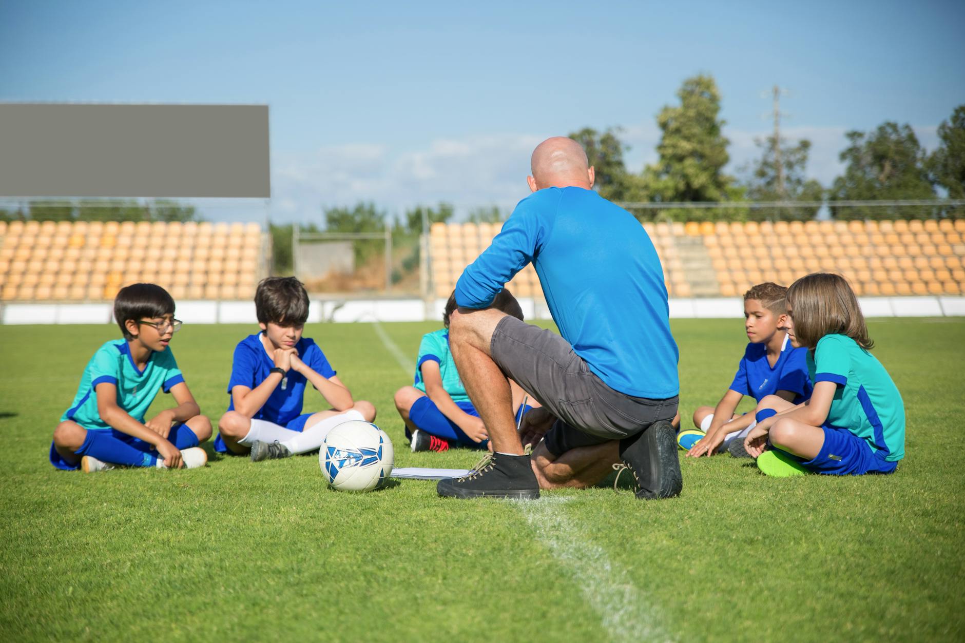 A Pro Touch Coach in blue shirt coaching a group of kids sitting on a football pitch